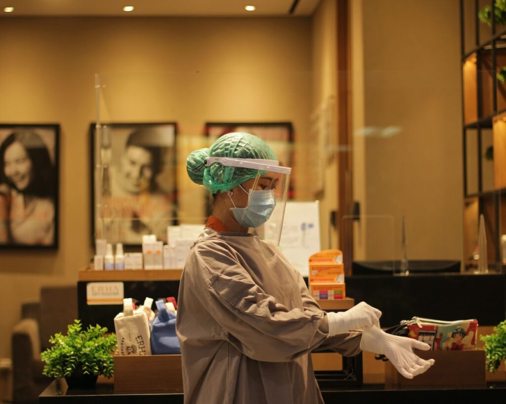 A health care professional puts on white nitrile gloves while wearing scrubs, a hairnet, and a face shield