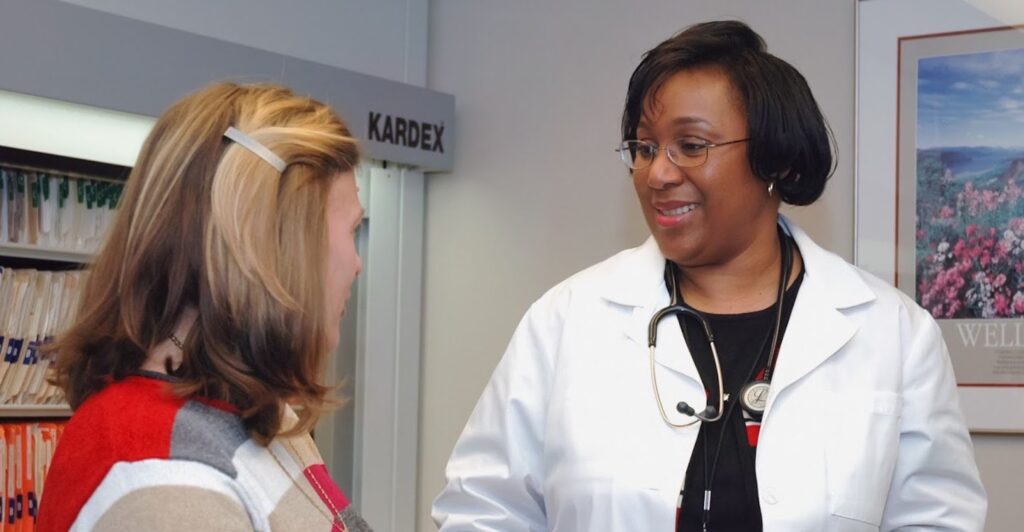 A female medical professional wearing a white lab coat has a conversation with a woman wearing a striped shirt.