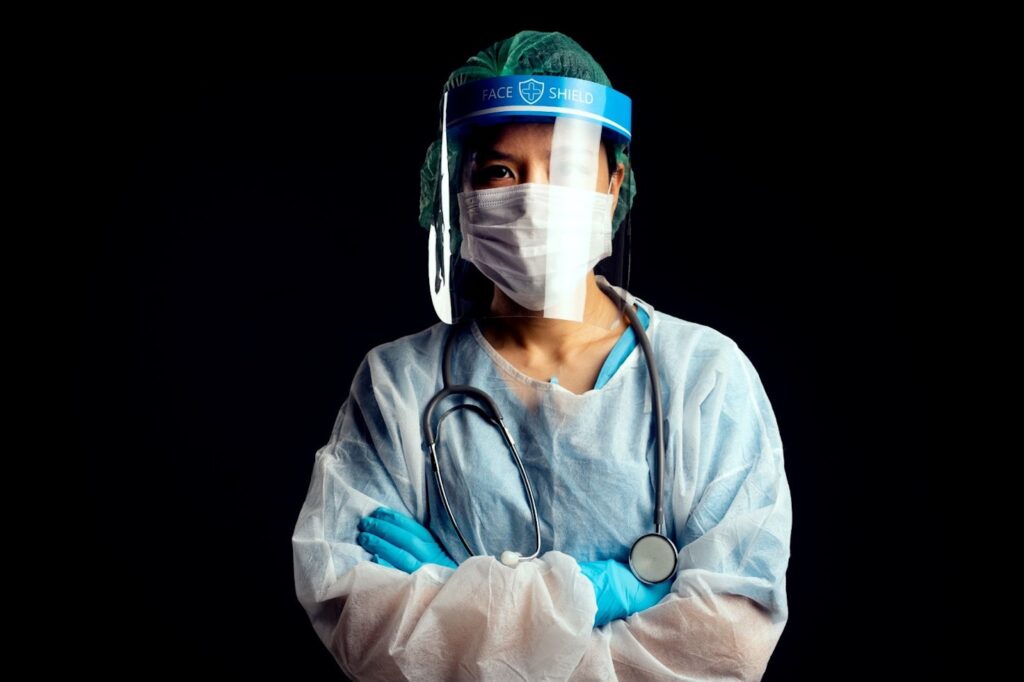 A medical professional stands with her arms crossed while wearing a face shield and medical gear.
