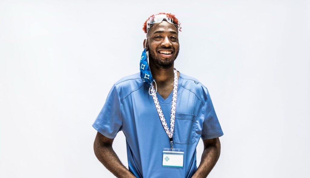 A male medical professional smiles while wearing blue scrubs and a face mask dangling off his ear.