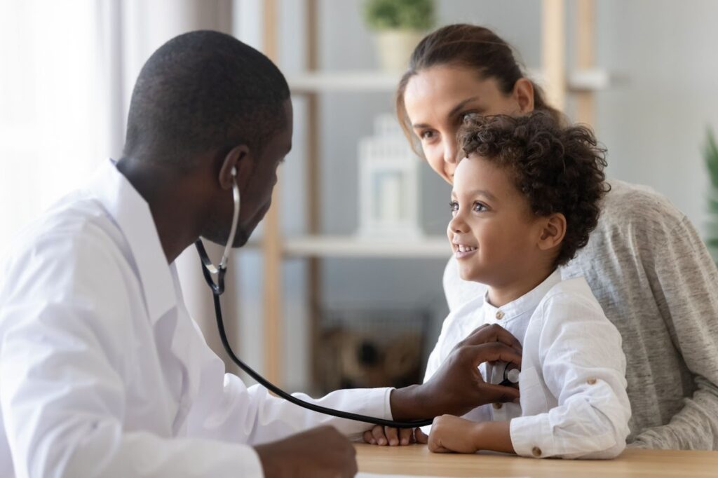 A medical professional holds a stethoscope to a young child's chest who is sitting on a woman's lap.
