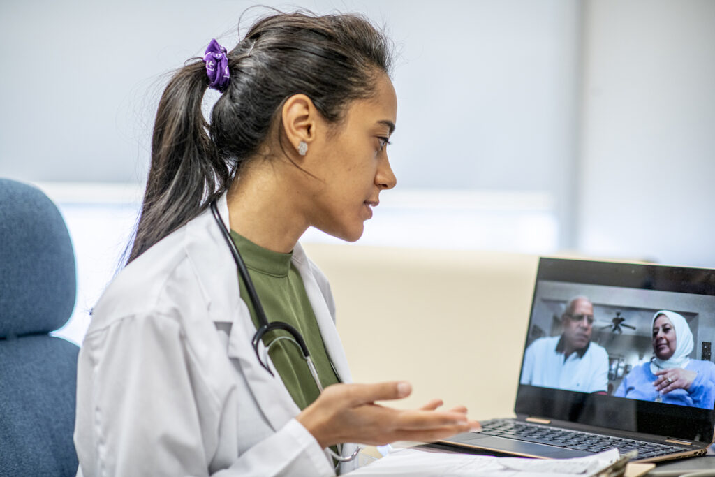 A physician assistant in a white lab coat speaks to two patients via video chat.