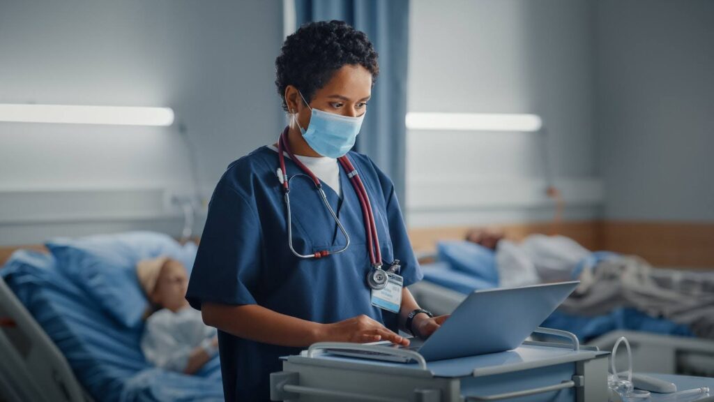 A hospital physician assistant at a patient's bedside enters notes into a laptop.