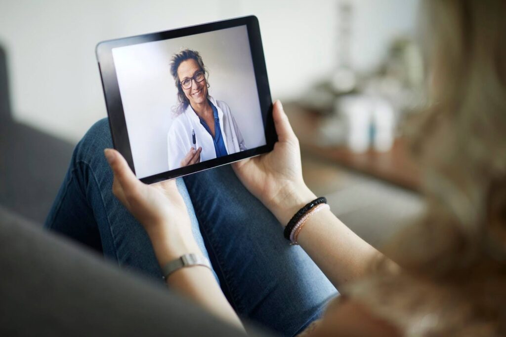 A patient holds a tablet; on its screen, we see a physician assistant delivering telehealth.