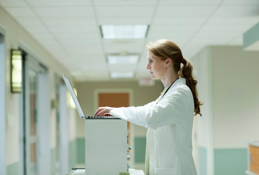 A physician assistant stands at a workstation, entering patient data on a laptop computer.